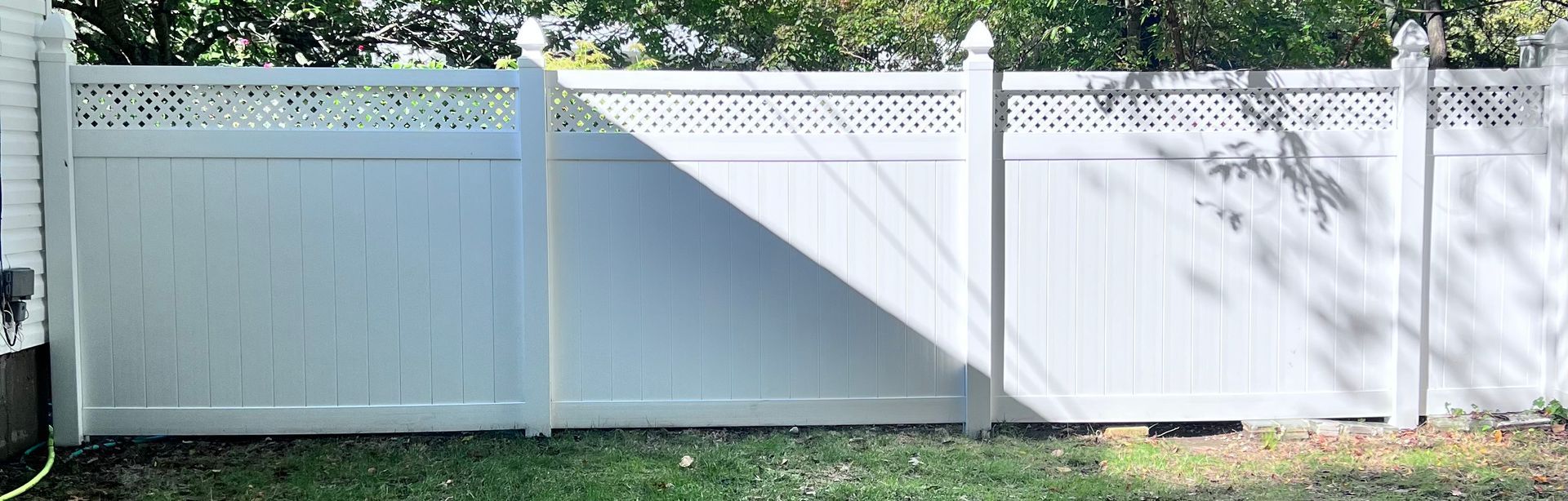 White vinyl fence with lattice top section, on grass in front of a house. Shadow of trees cast across the fence.