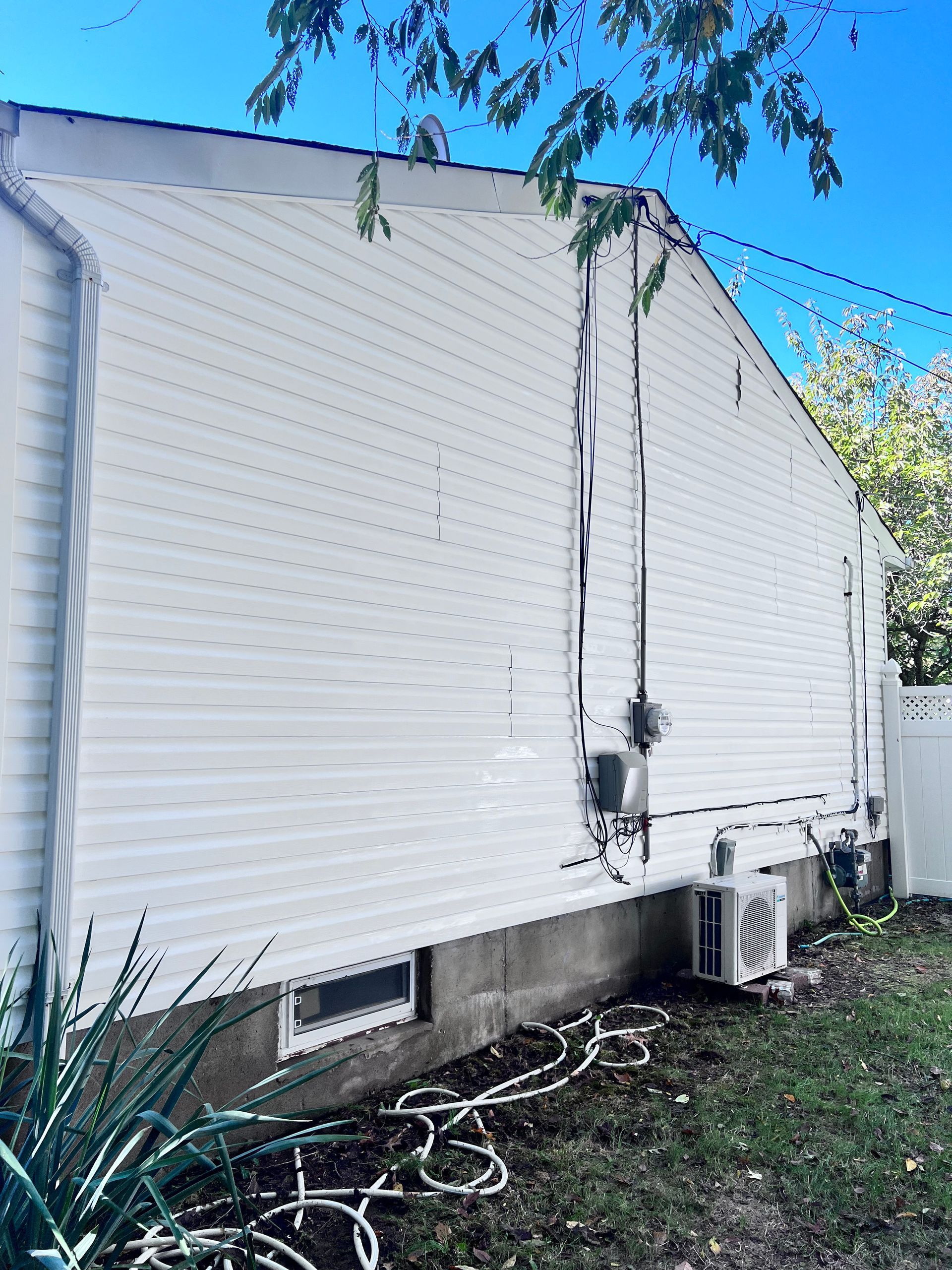 Side view of a white-sided house with a gas meter, air conditioning unit, and electrical lines. A small window is visible near the base.