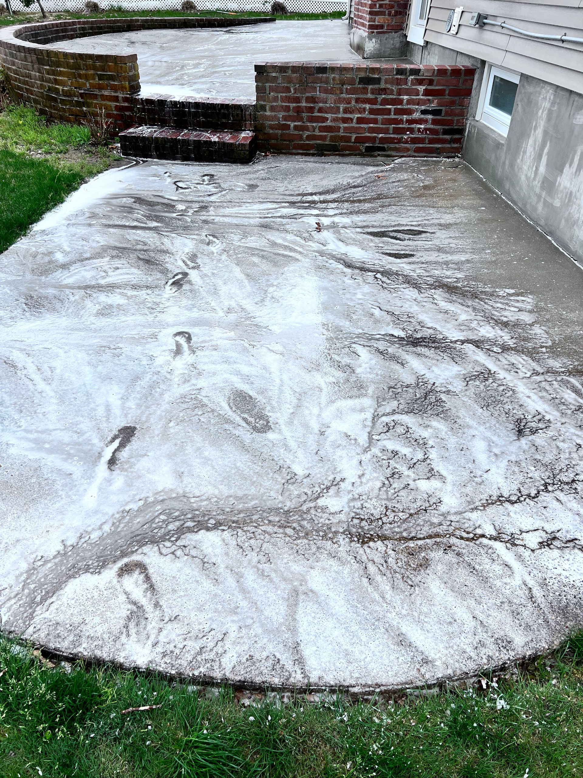Concrete patio covered in foamy cleaning solution; surrounded by grass and brickwork.