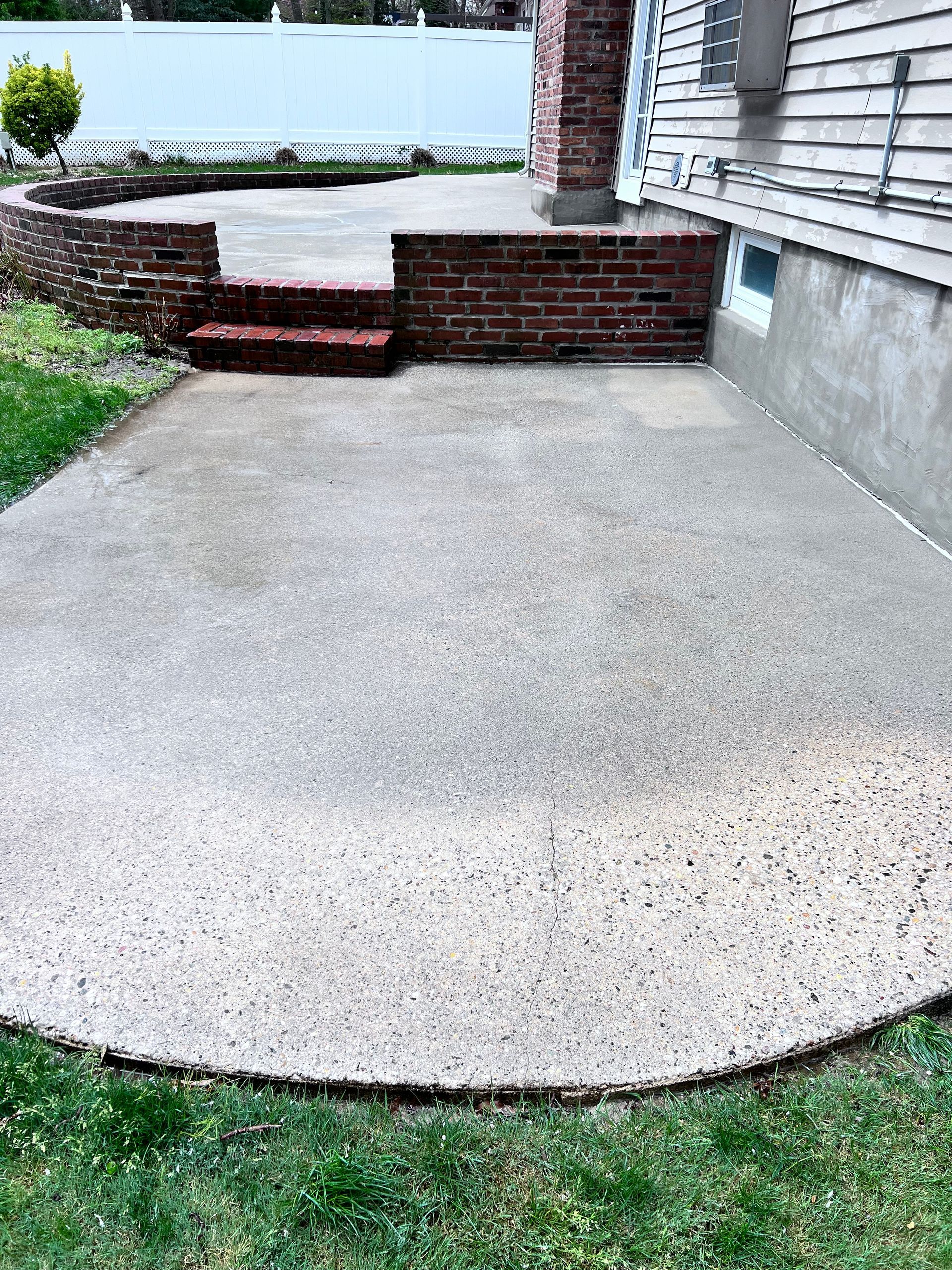 Concrete patio bordered by grass and brick. A small brick wall and steps lead to a higher level.  A chimney and house siding are to the right.