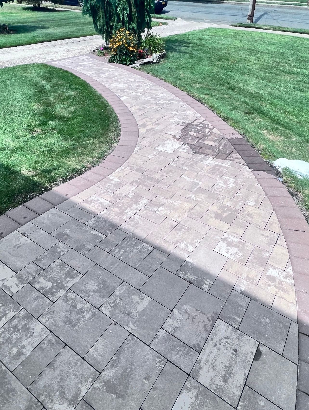 Curving paved walkway with red brick borders through green grass, leading towards a small flower bed and street.