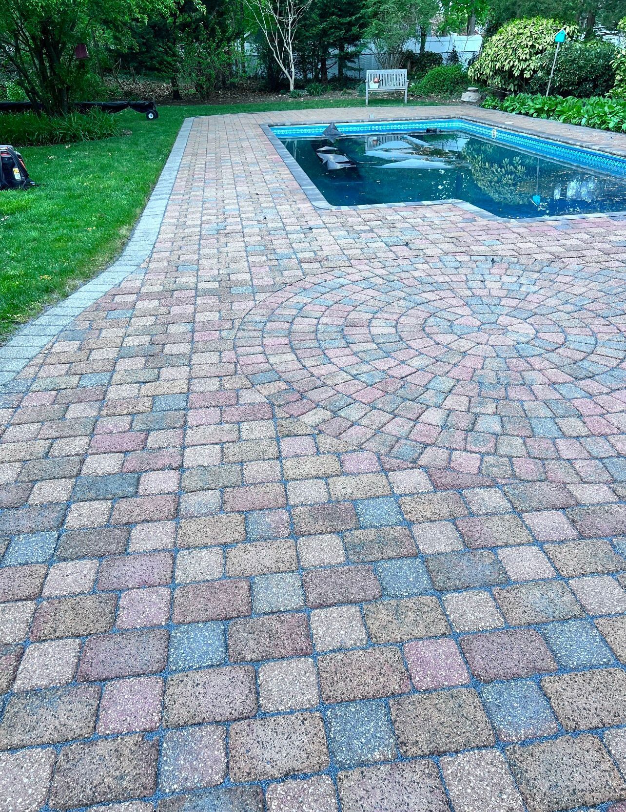 Brick patio surrounds a pool with blue water, surrounded by green grass and trees.