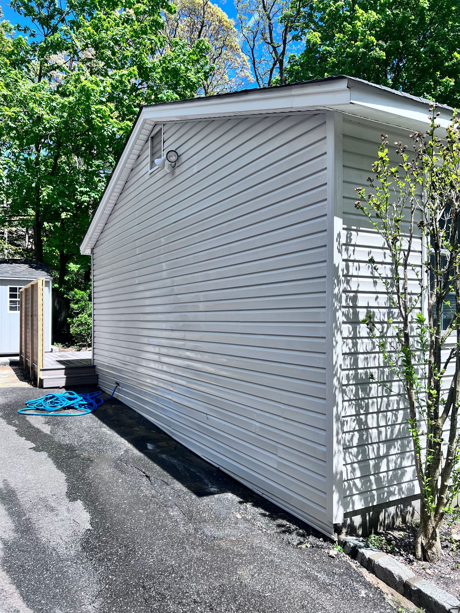 Side view of a light gray building with horizontal siding, next to a paved driveway, surrounded by trees.