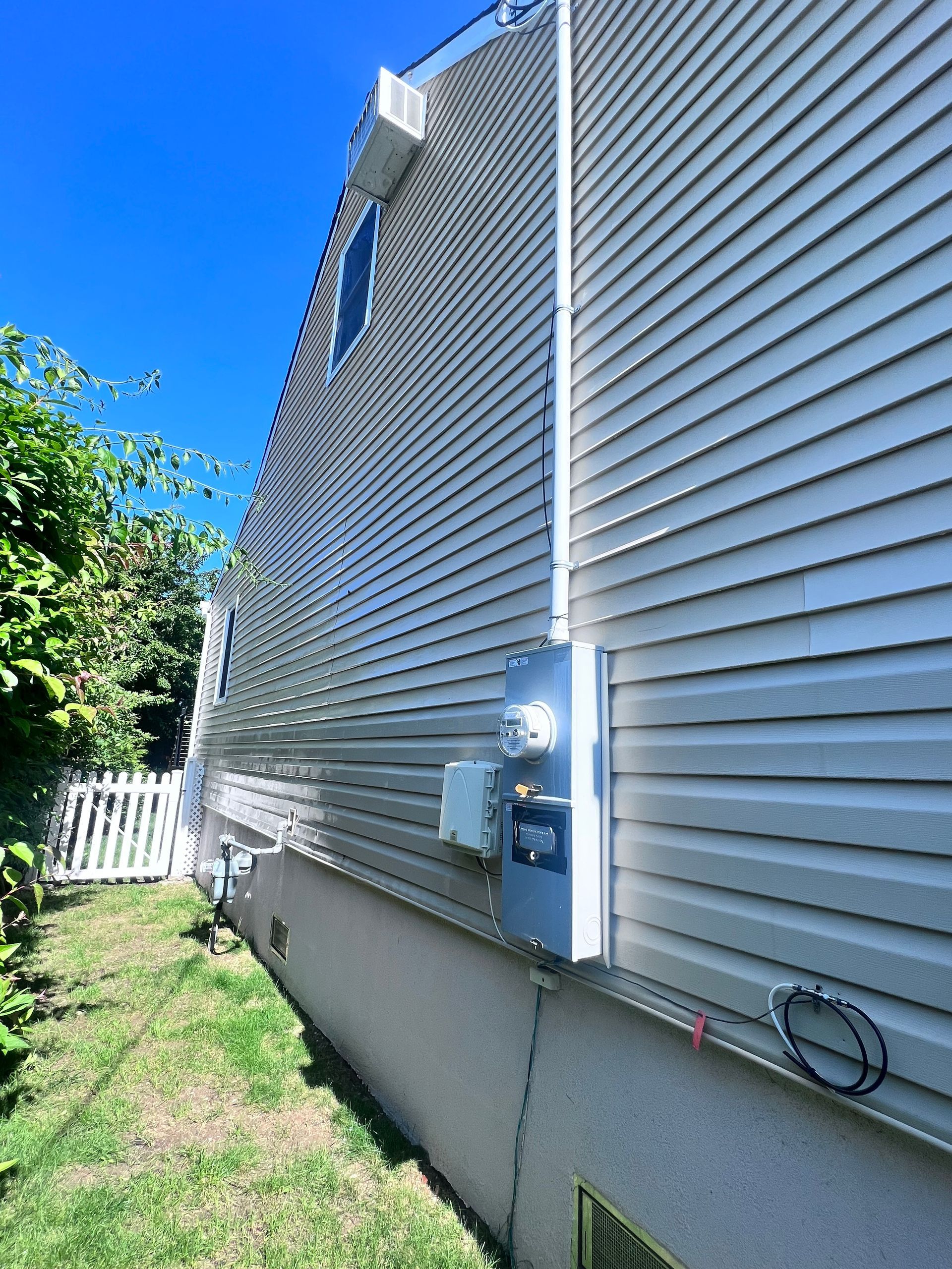 Side view of a house with gray siding, a white fence, and an electrical meter box on the wall; sunny day.