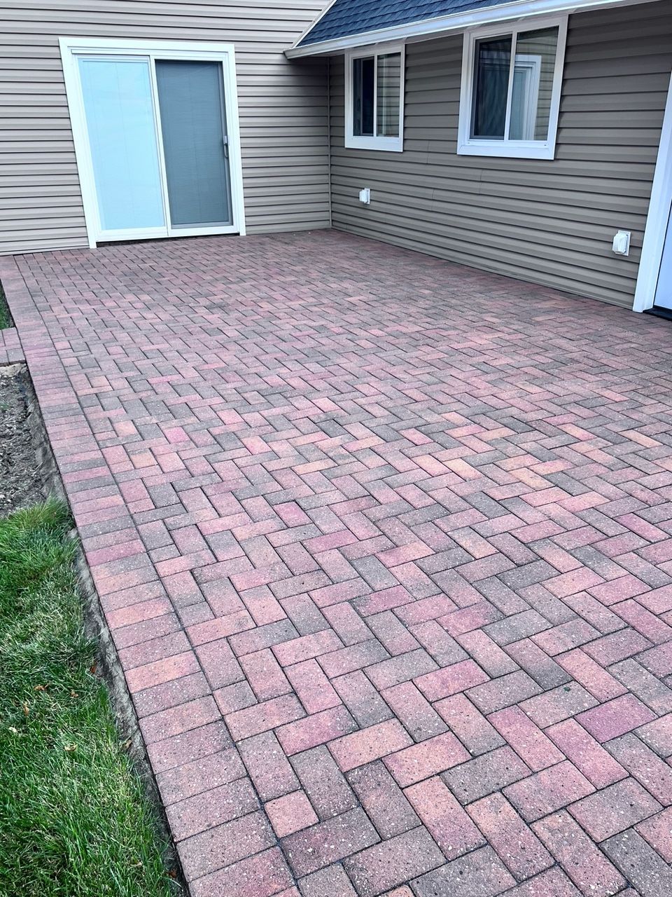 Brick patio next to a house with a sliding glass door and two windows; the patio's edge borders green grass.