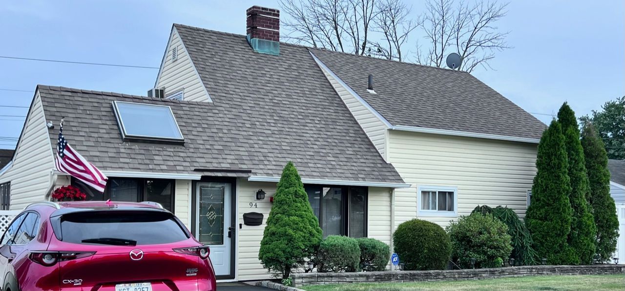 A house with a red car parked in front. An American flag hangs from the house. Evergreen bushes and trees are in the front yard.
