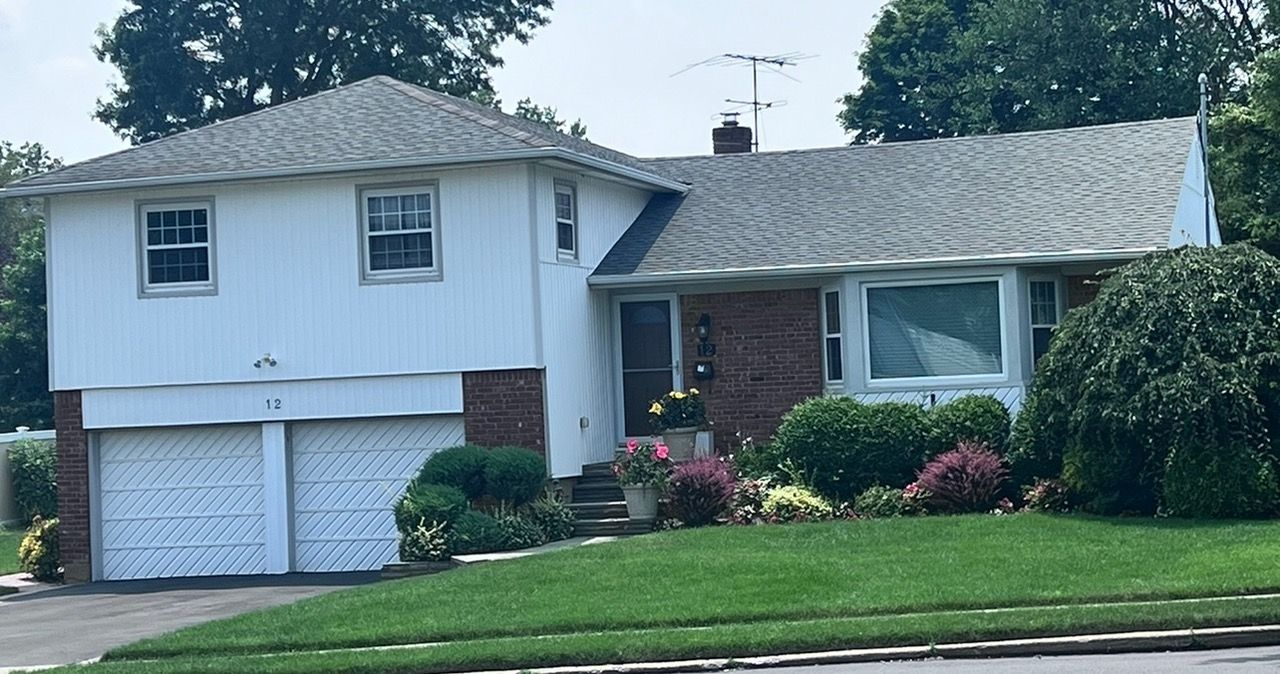 Two-story white house with a two-car garage, brick accents, and a well-manicured lawn. Bushes and flowers line the front.
