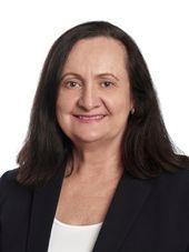 Woman with dark hair and a navy blazer smiles at the camera against a white background.