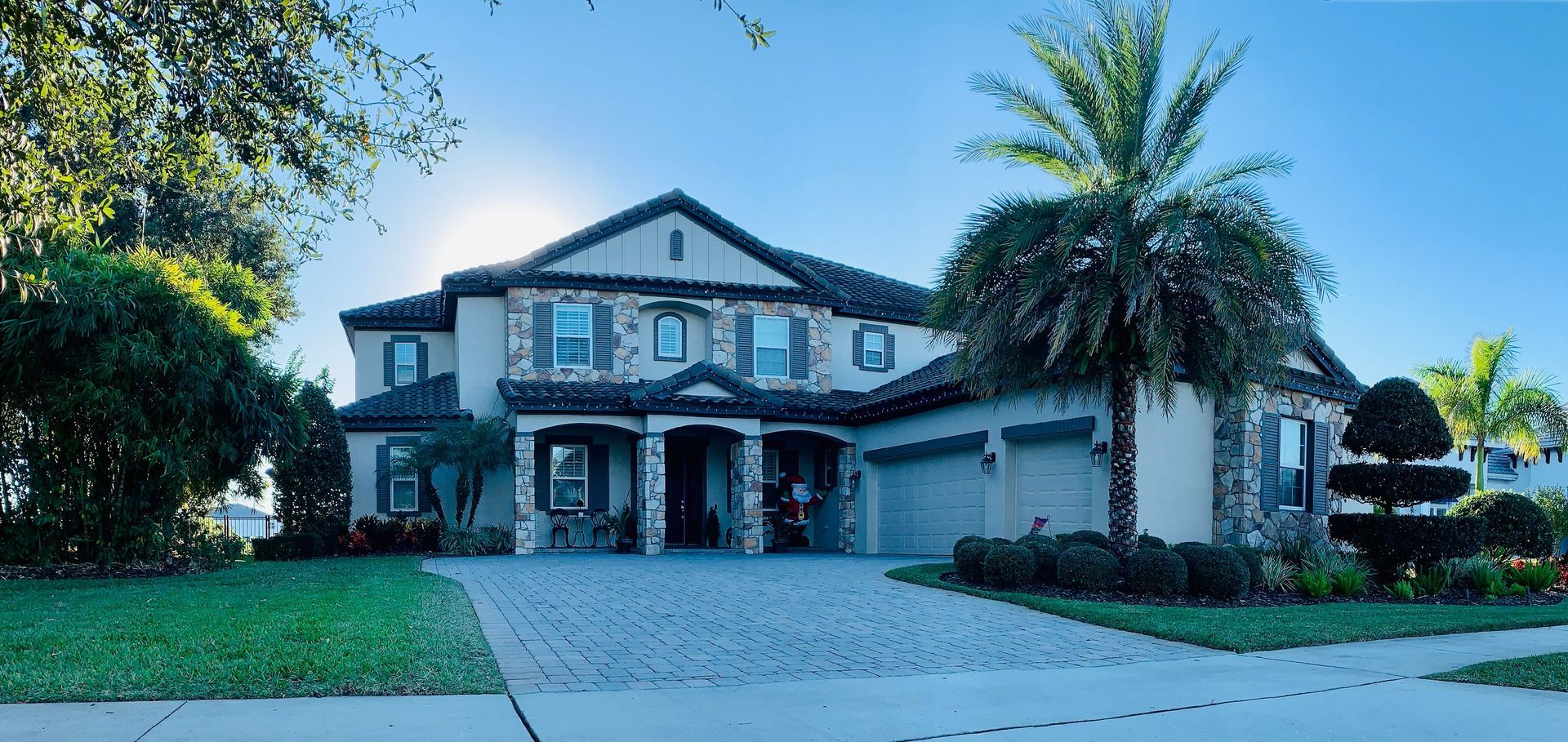 Large two-story house with a brick driveway, lawn, and palm trees under a bright blue sky.