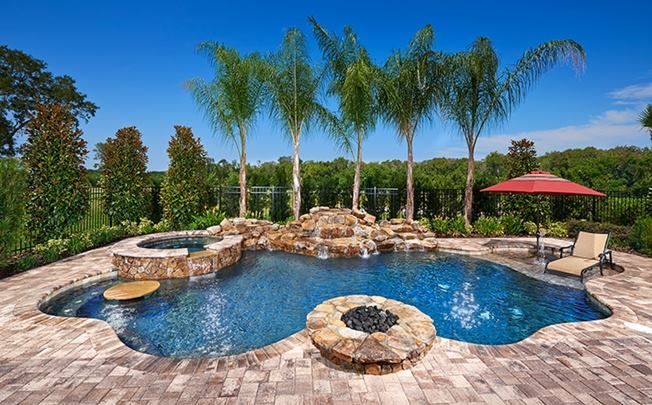 Swimming pool with waterfall feature, spa, and fire pit in a landscaped backyard under a blue sky.