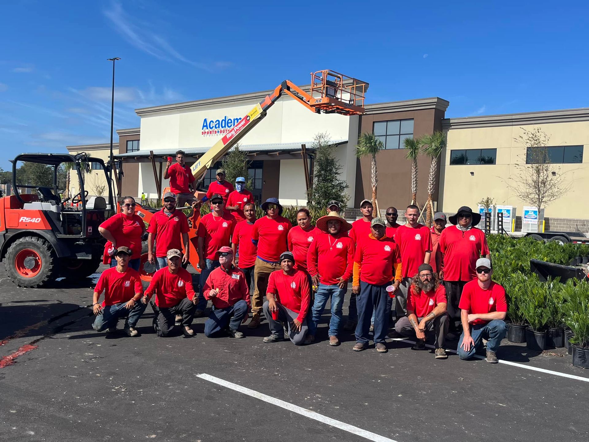 Group of people in red shirts pose in front of a building with a forklift and lift on a sunny day.