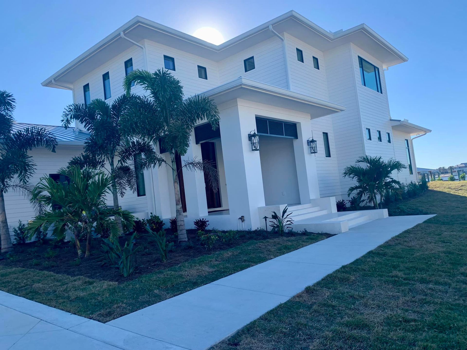 White two-story house with palm trees, blue sky. Concrete walkway, green grass, and sunlight.