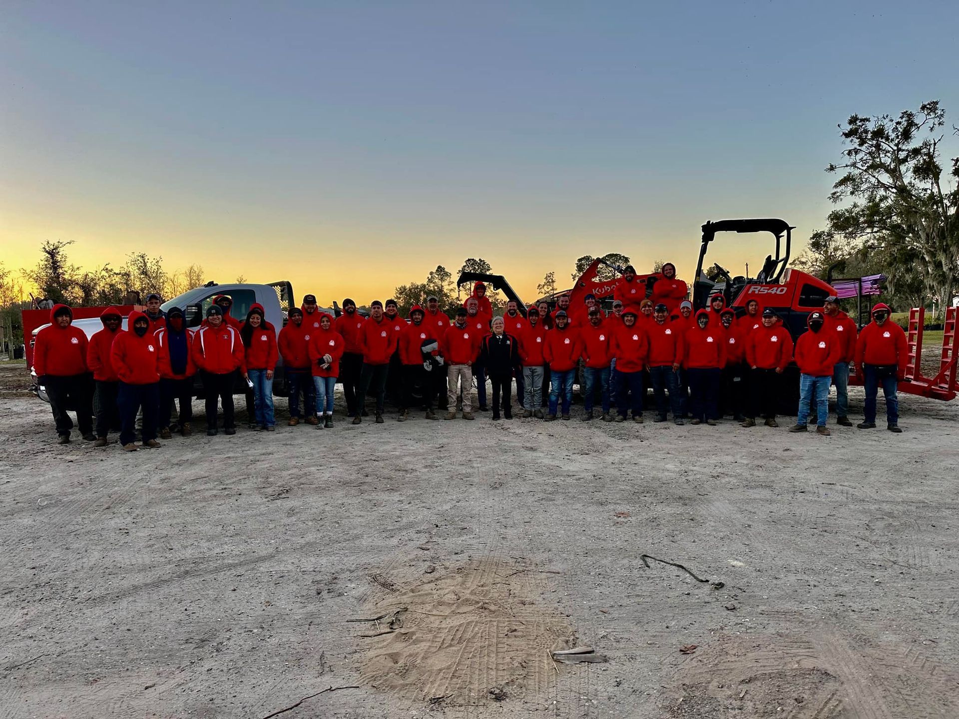 Group of people in red jackets gathered outdoors with trucks and a tractor at dusk.