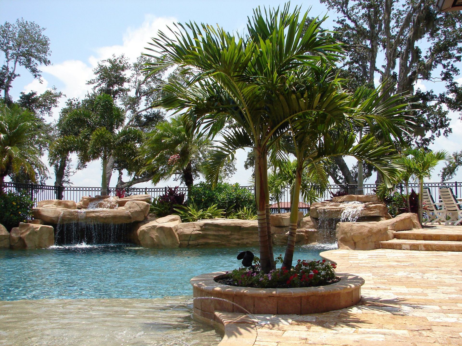 Palm trees next to a pool with waterfalls and a stone patio on a sunny day.