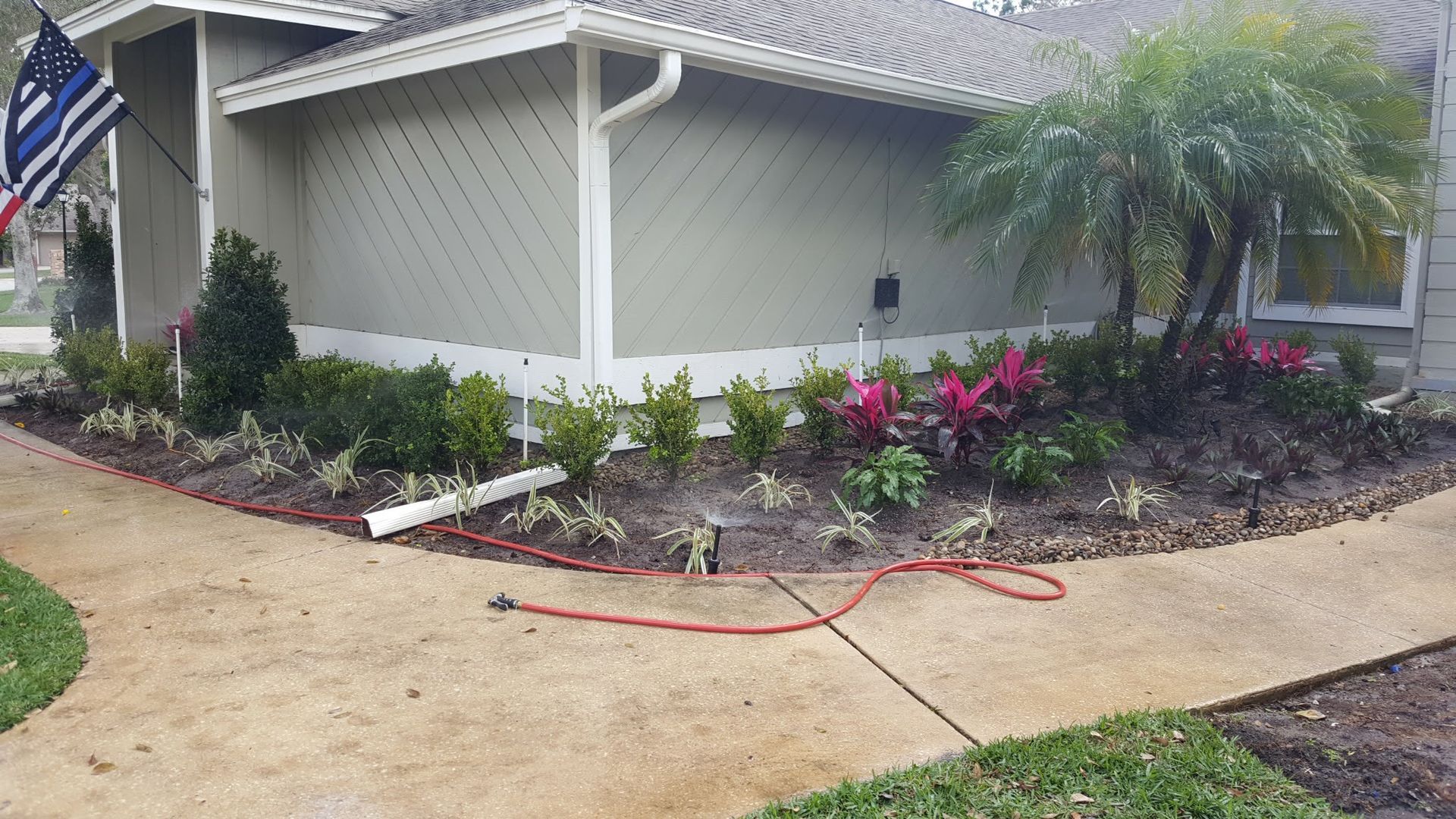 House with landscaped garden bed, red hose on path, green grass, American flag.