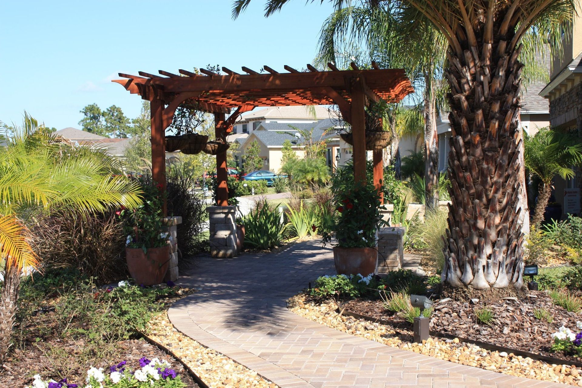 A wooden arbor frames a brick pathway in a garden, with flowerbeds and palm trees.