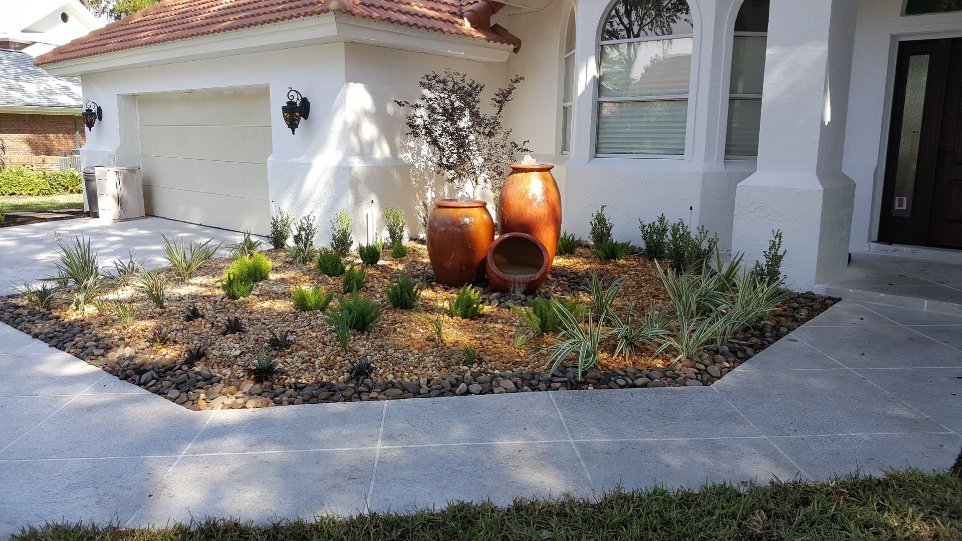 Landscaped front yard with potted plants and pots, in front of white house with garage.