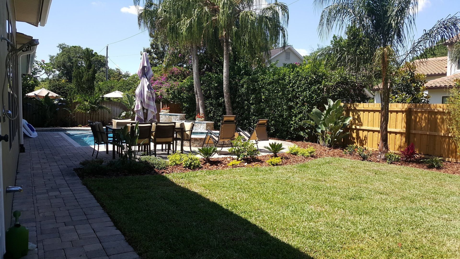 Backyard with pool, patio furniture, green grass, and trees under a sunny sky.