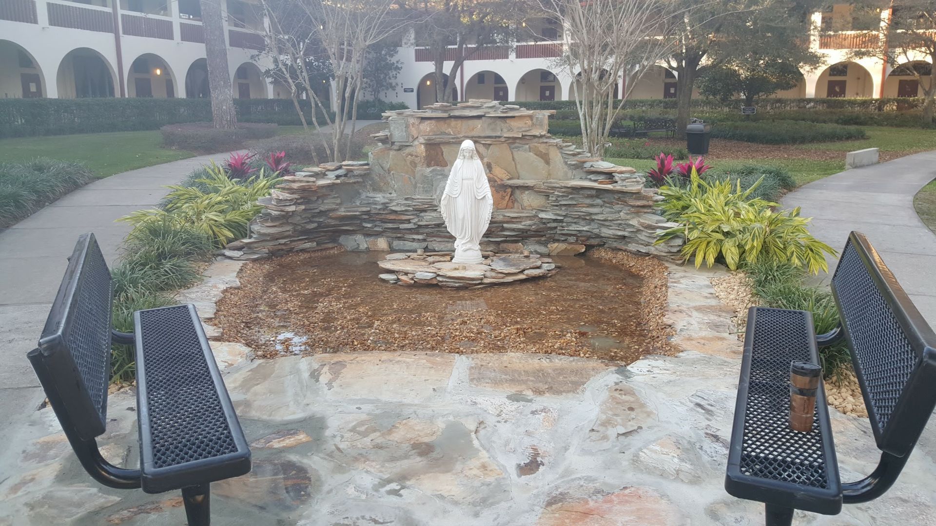 Fountain with Virgin Mary statue, stone benches, and arched buildings in the background.