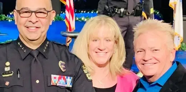 Three people smiling at the camera, indoors. The man on the left wears a police uniform. The woman in the middle wears a pink blazer. The man on the right wears a blue shirt.