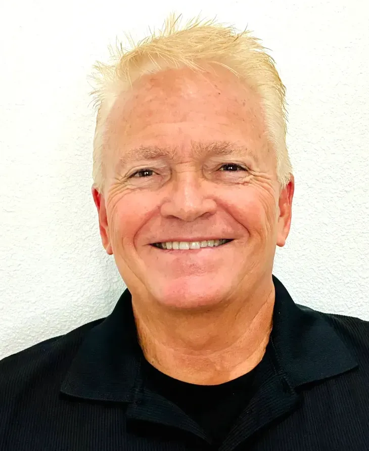 Smiling man with short, spiky blond hair, wearing a black shirt, against a white background.