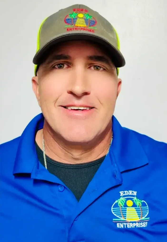 Man wearing a hat and blue shirt smiles at the camera. He's inside, against a light gray wall.