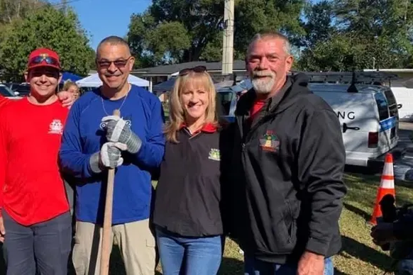 Four people stand outside, smiling. The man on the right holds a shovel.