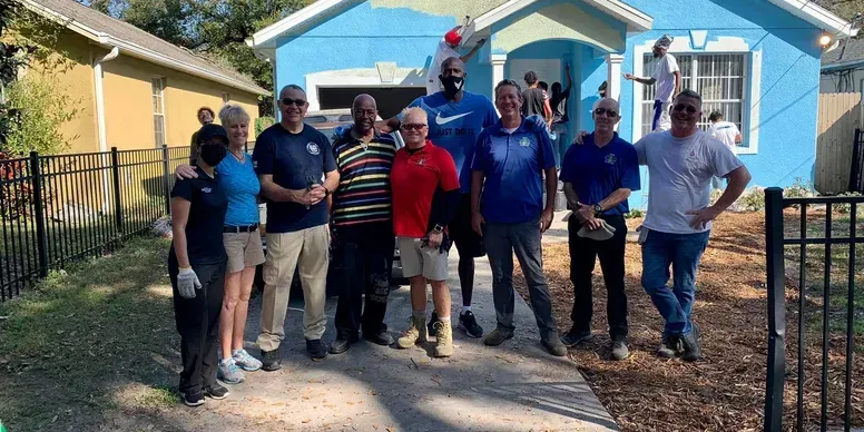 Group of people standing in front of a blue house, some wearing work attire.