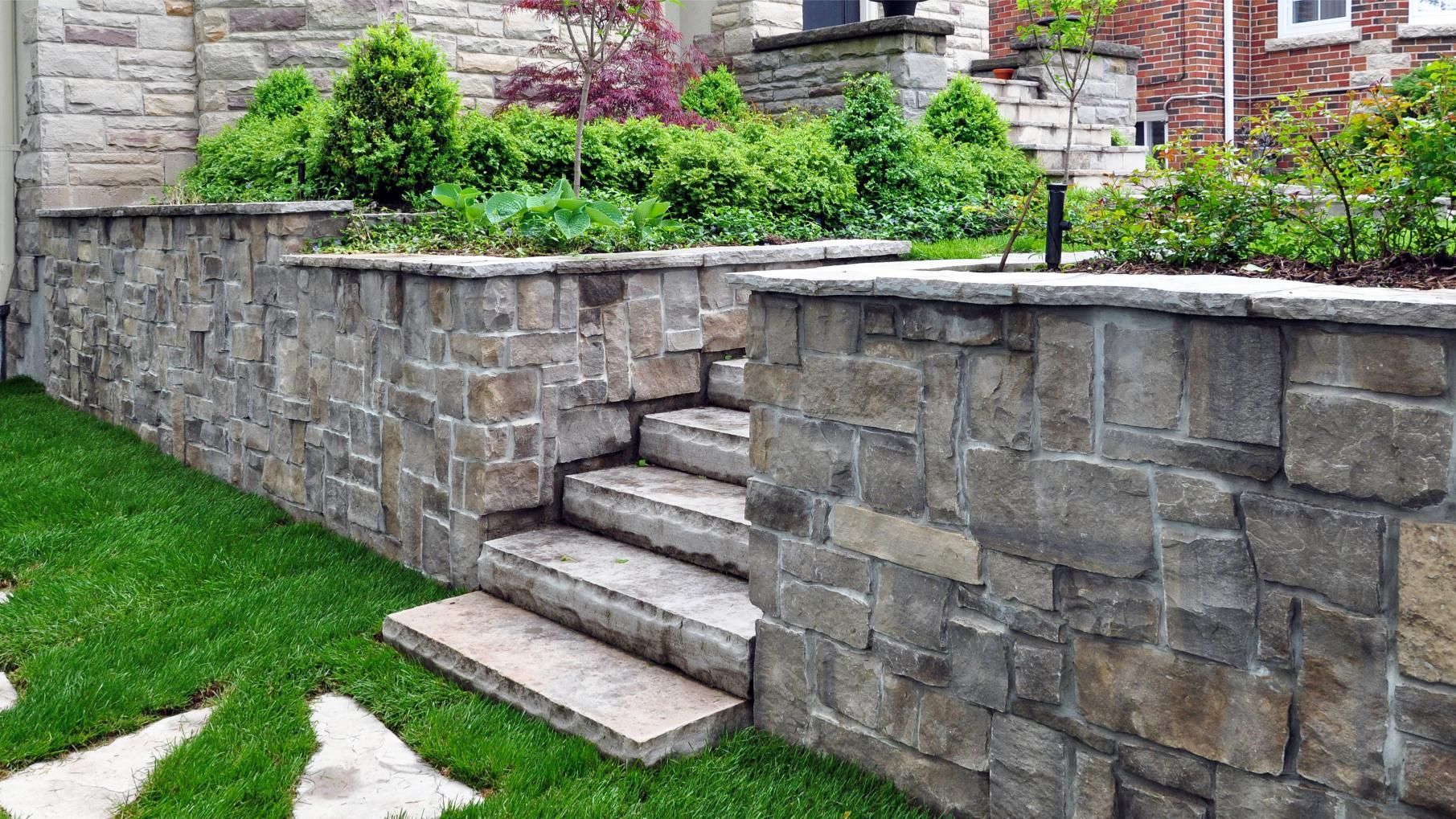 A stone retaining wall with integrated steps leading to a raised garden bed filled with green shrubs and trees.
