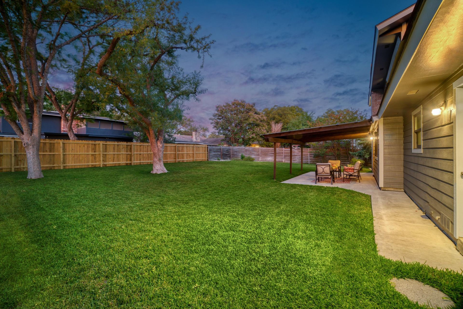 A grassy backyard at dusk with a wooden fence, large trees, and a patio covered by a small roof attached to a gray house.