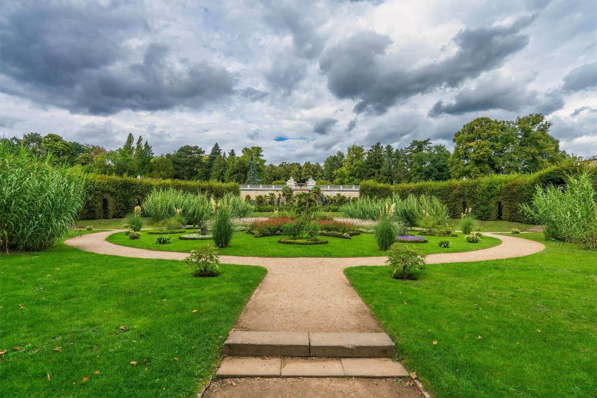 A garden with a gravel path leading to a circular lawn surrounded by hedges and trees under a cloudy sky.