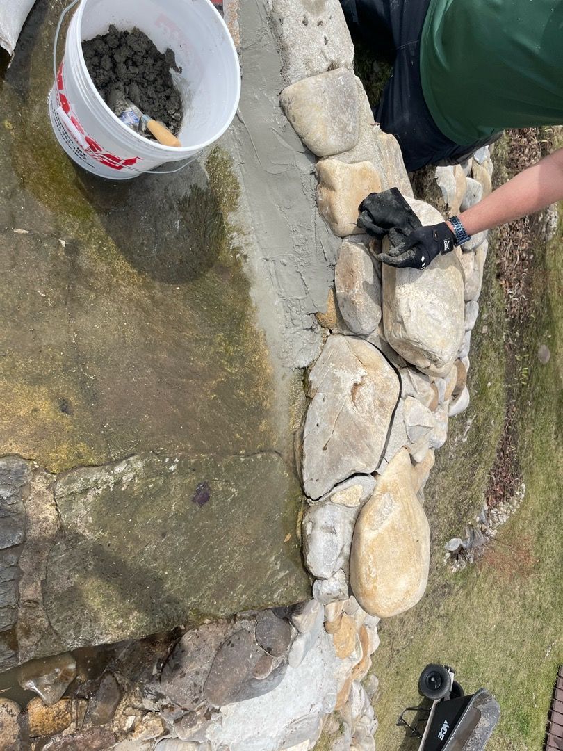 Person repairing a stone structure with mortar and a bucket nearby.