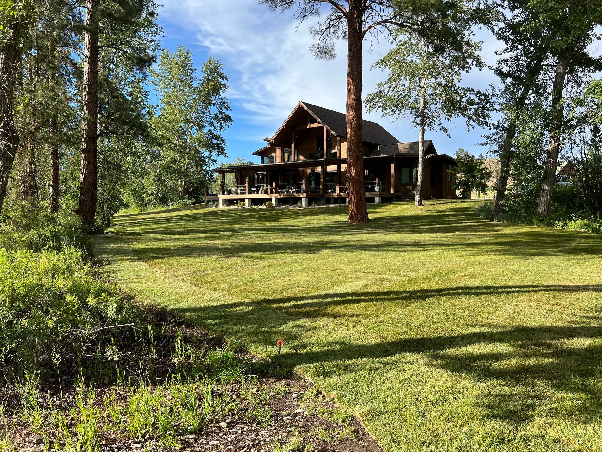 Large wooden house with porch on a grassy lawn, surrounded by trees under a blue sky.