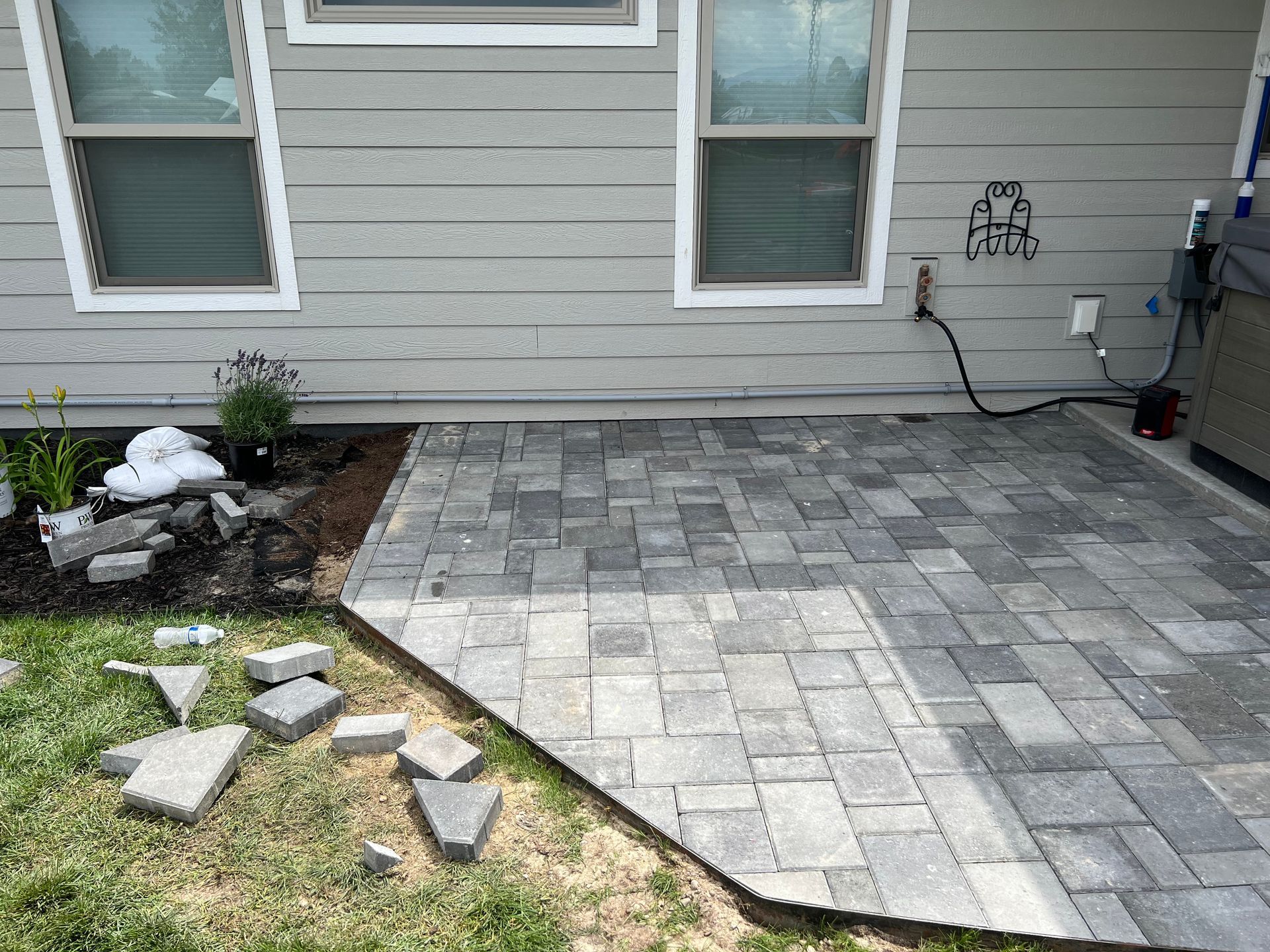 Gray brick patio under construction next to a house with two windows; some bricks are in a pile on the grass.