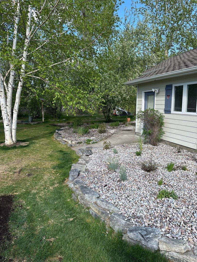 A house with a stone-edged flower bed filled with plants and gravel, beside a green lawn and trees.