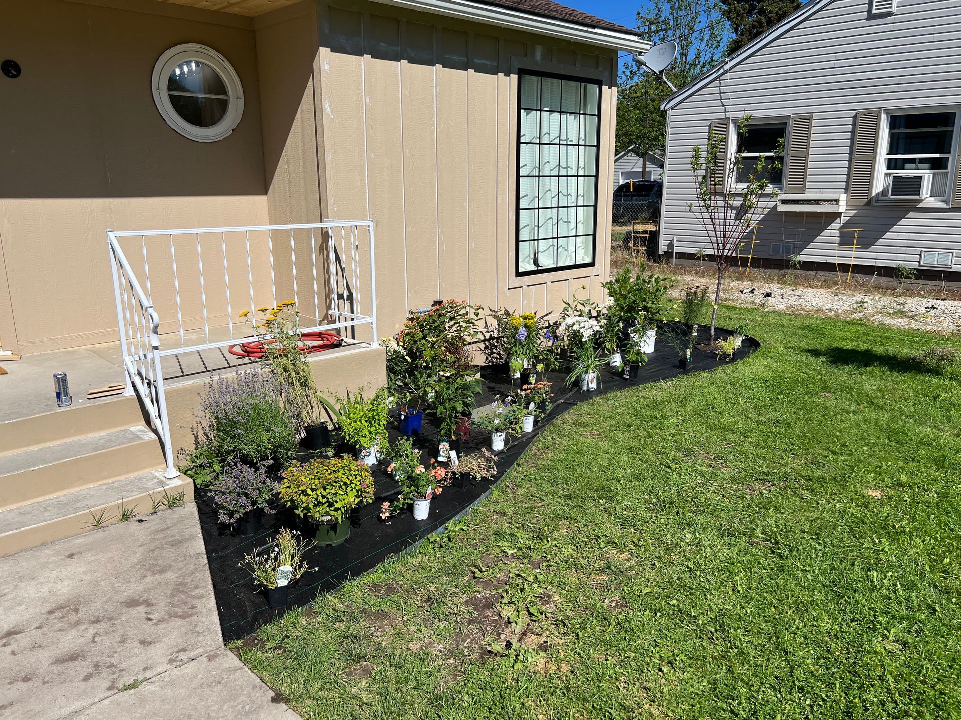 Tan house with flower bed, white railing, and a window; green lawn and partial view of a neighboring house.