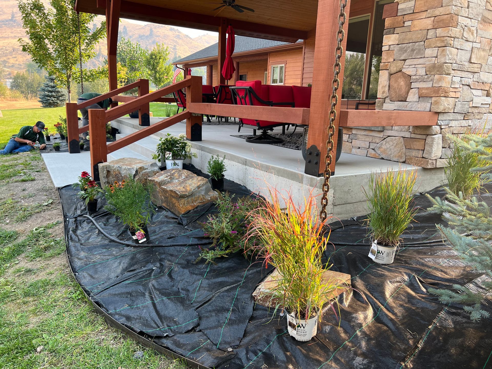Landscaping project: black fabric, plants, rocks, porch with red furniture, stone wall, person working.