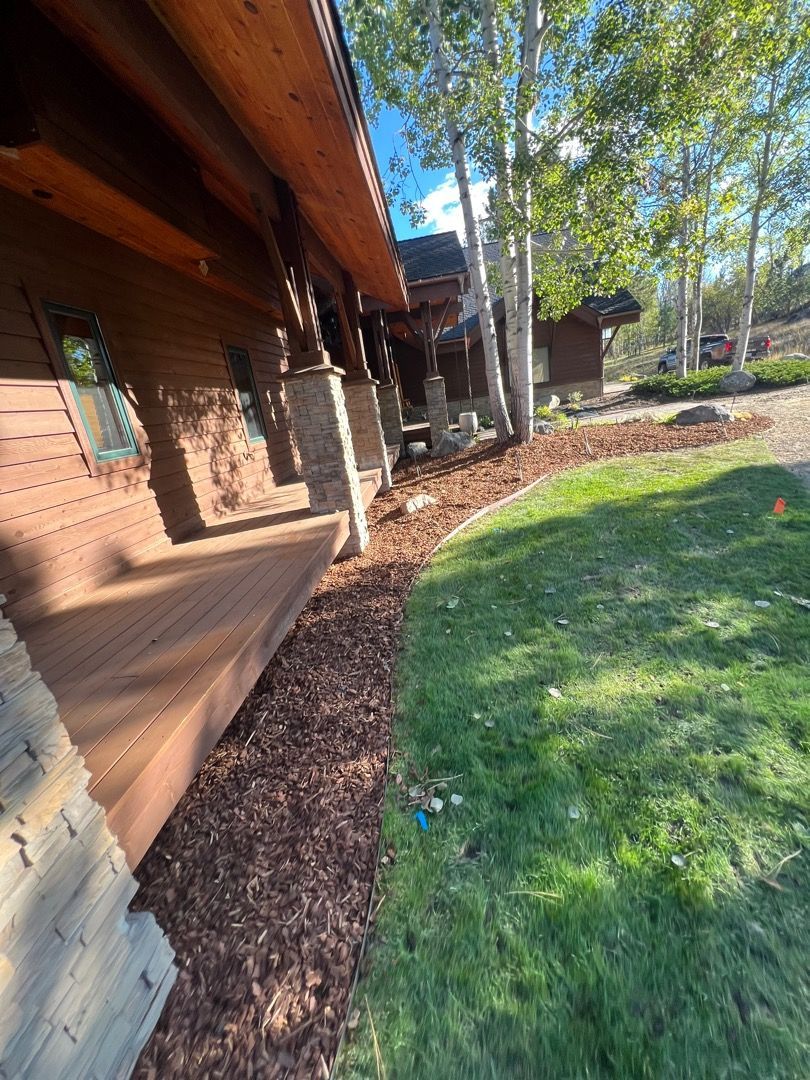 Wooden house with porch and mulch bed next to grass and trees.