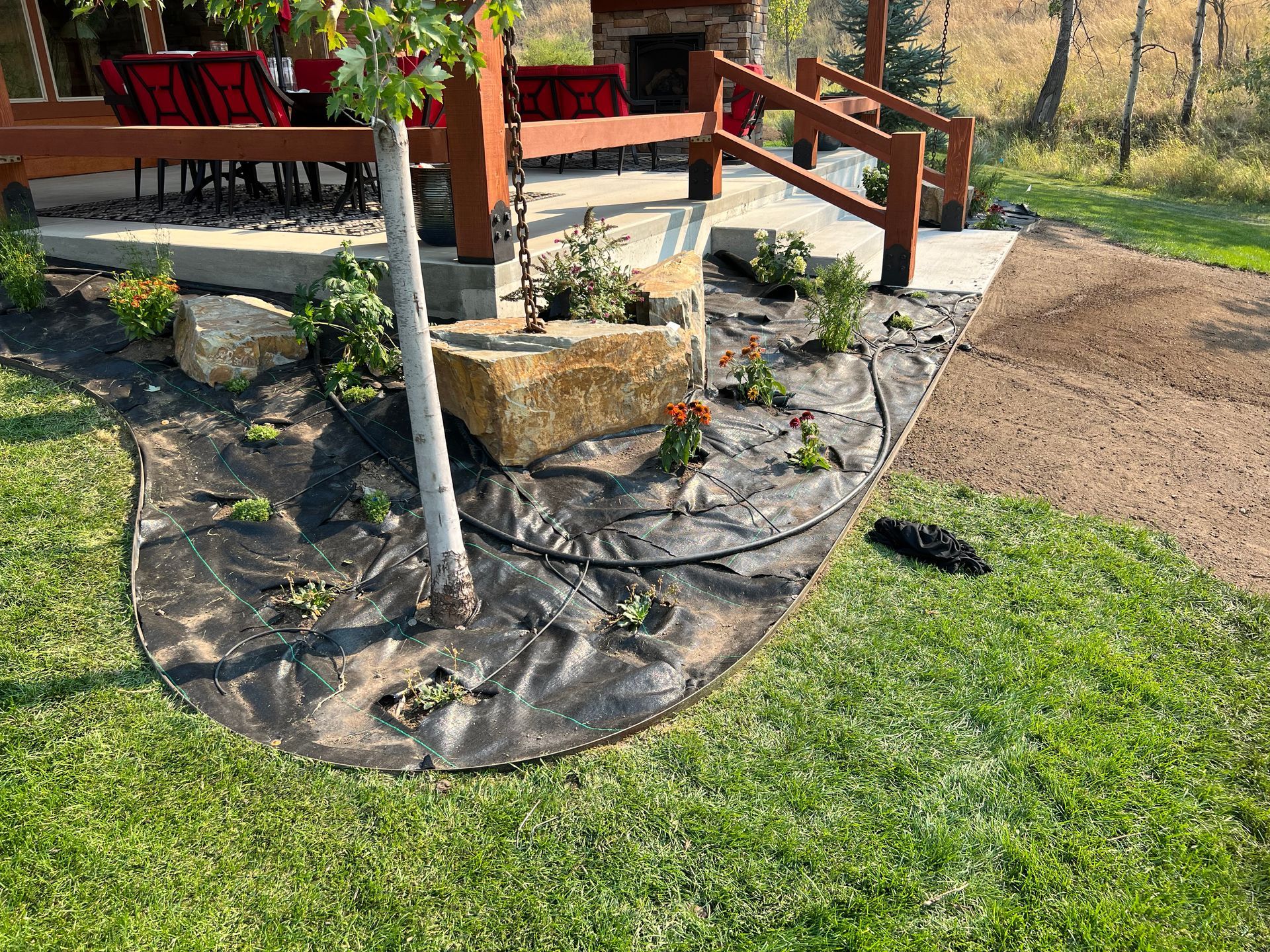 A landscaped garden bed with dark fabric, large rocks, and small plants next to a wooden deck and walkway.