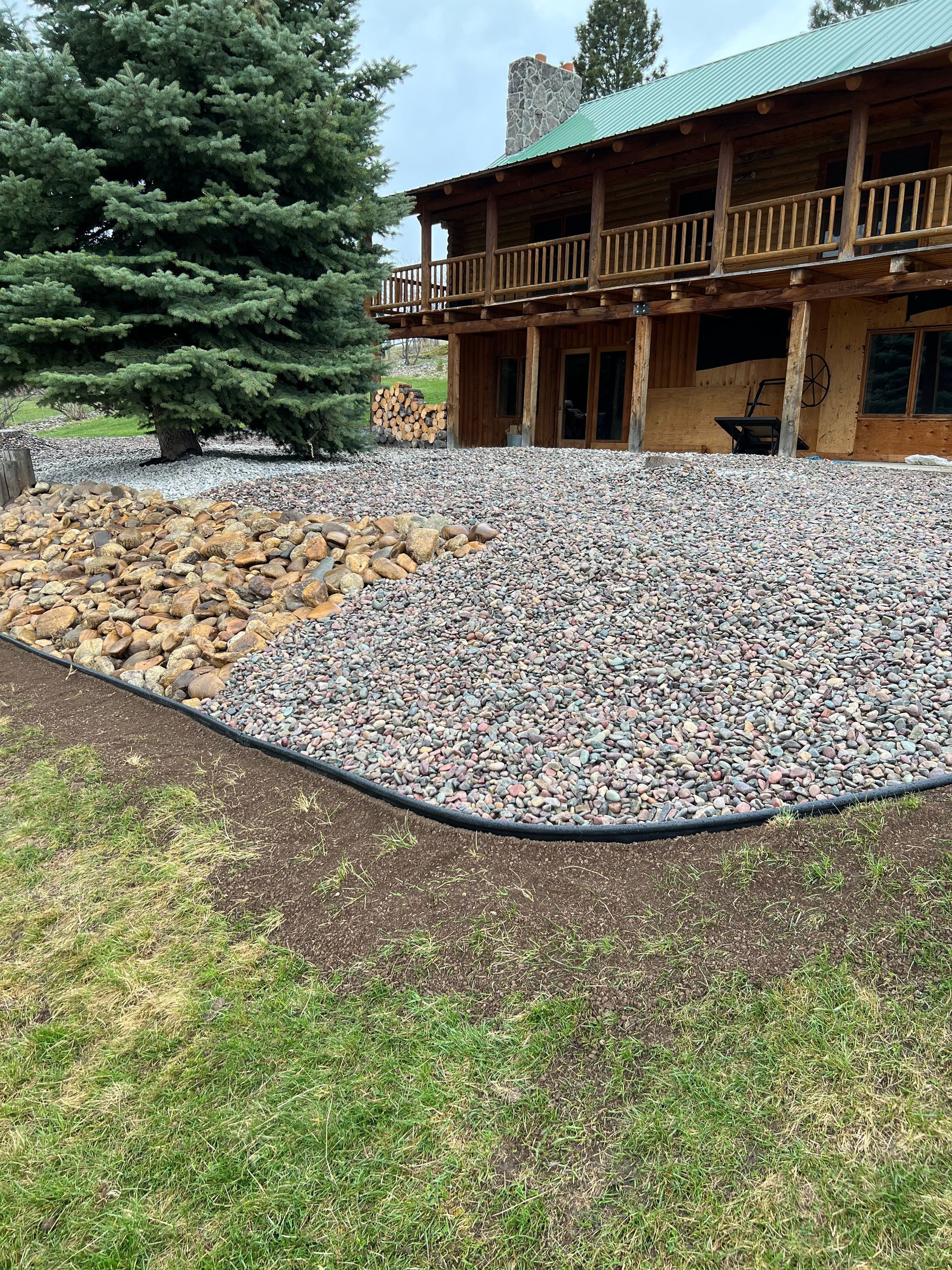 Rock garden with a wooden house in the background and a large evergreen tree to the left.