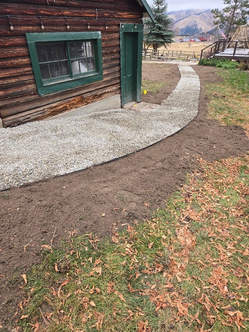 A gravel pathway curves alongside a rustic cabin with a green door and window. Dirt and grass surround it.