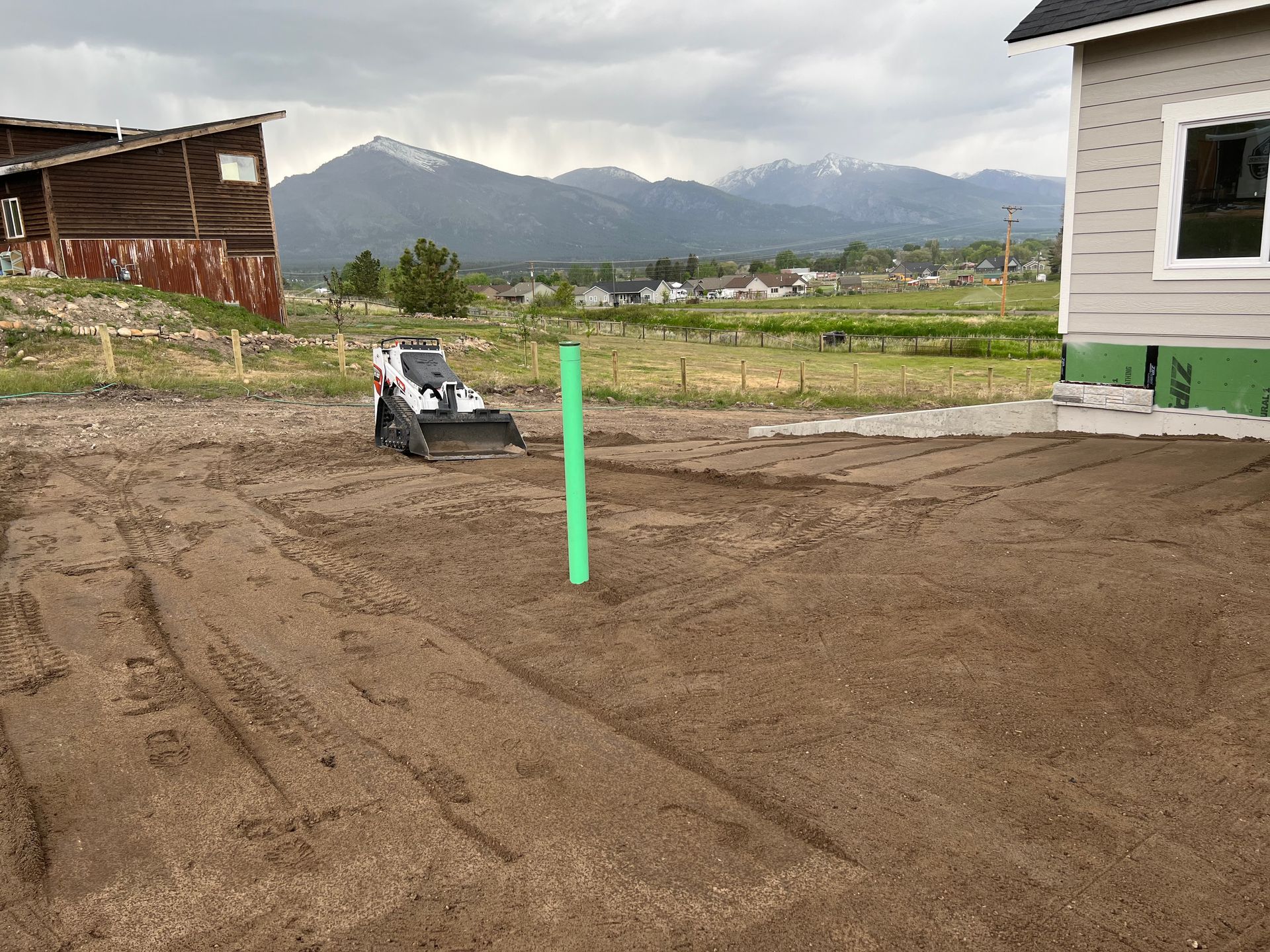 Bobcat on a dirt lot with a green pipe, a house, and a barn in a mountainous rural setting.