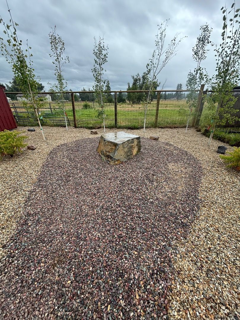 Rock fountain in a gravel bed, with birch trees and a fence in the background under a cloudy sky.