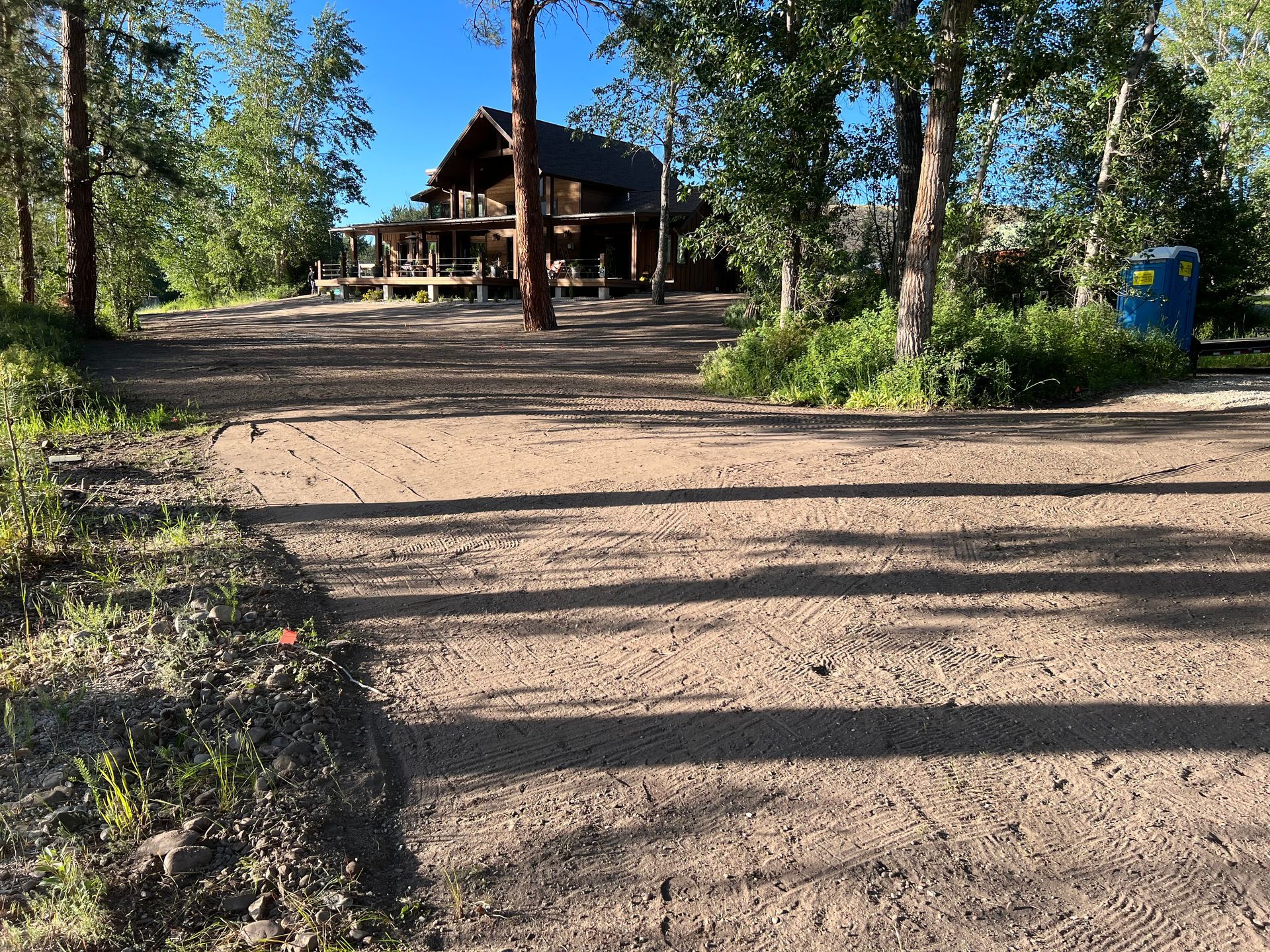 Dirt road leading to a wooden cabin surrounded by trees under a blue sky.