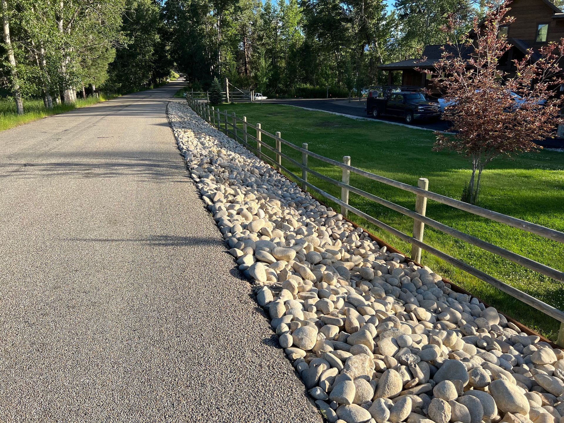 Gravel driveway bordered by large rocks and a wooden fence, leading towards trees and a house.