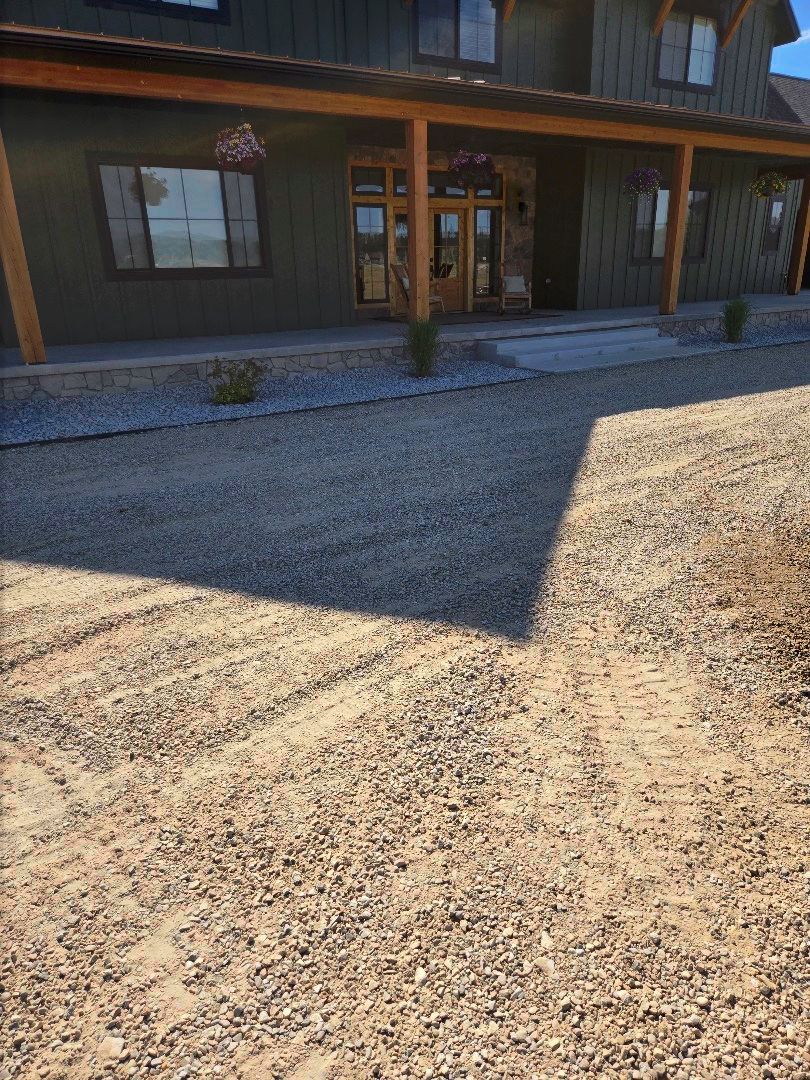 Gravel driveway leading to a green building with a wooden porch and windows. Sunlight casts a large shadow.