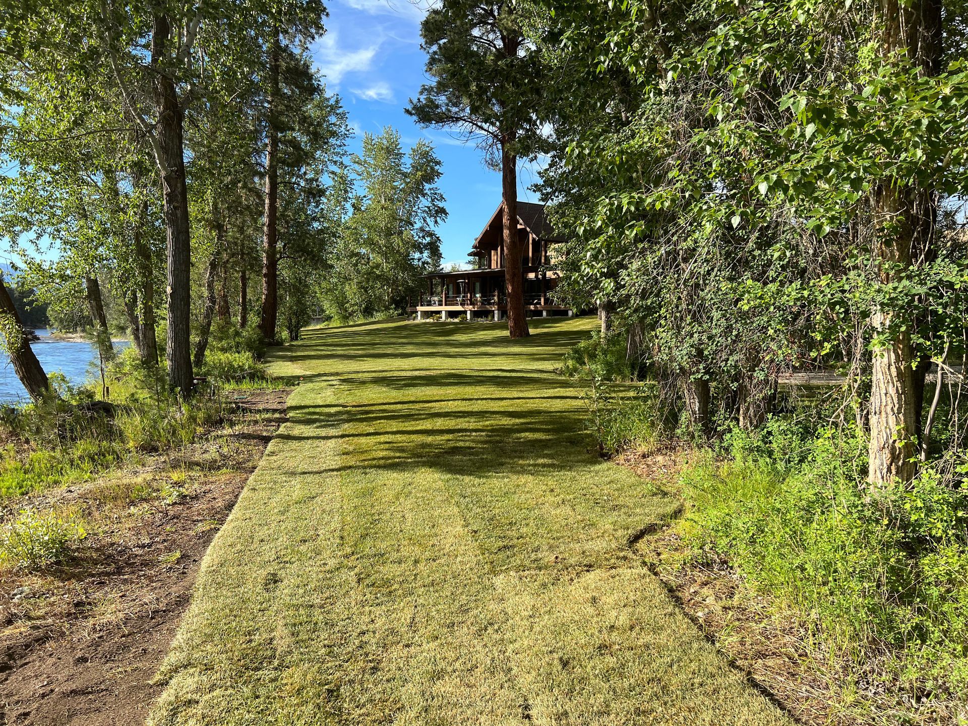 A grassy path leads to a house near a lake, framed by trees under a blue sky.