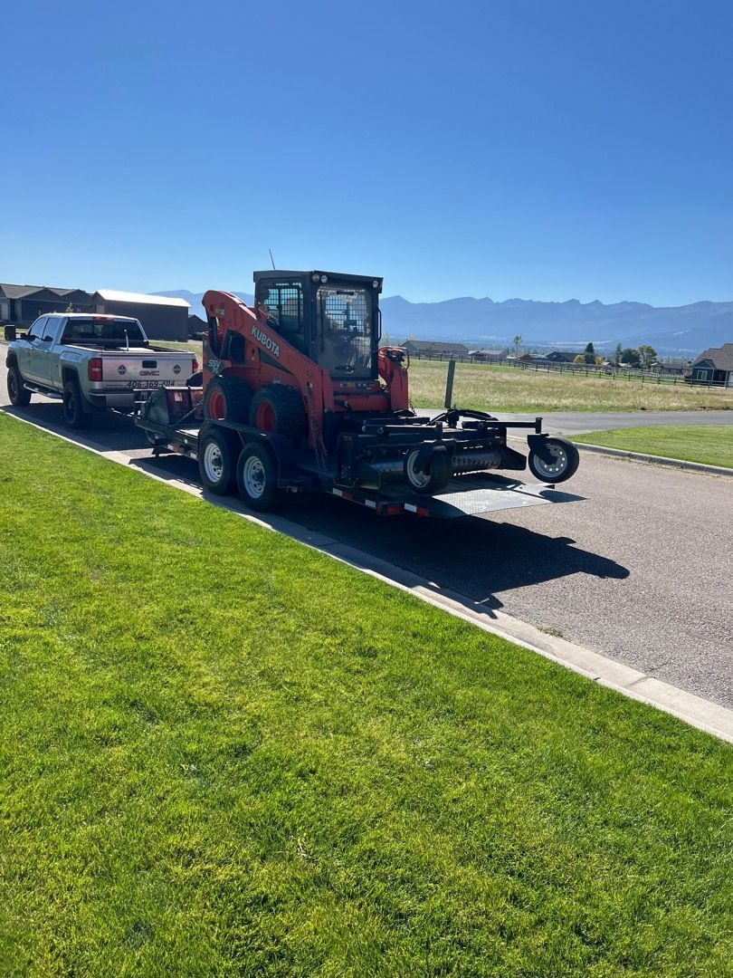Red skid steer loader on trailer, towed by pickup truck on residential street, sunny day.