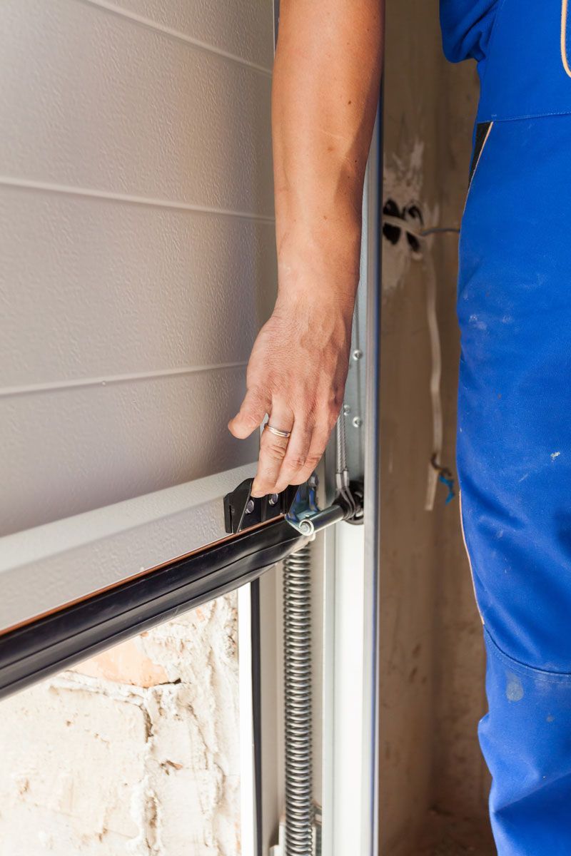 Person in blue overalls adjusting a garage door's mechanism, focusing on the hinges and spring.