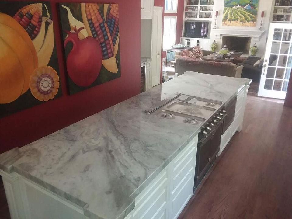 Kitchen island with marble countertop, stove, and white cabinetry, red wall with artwork, wooden floor.